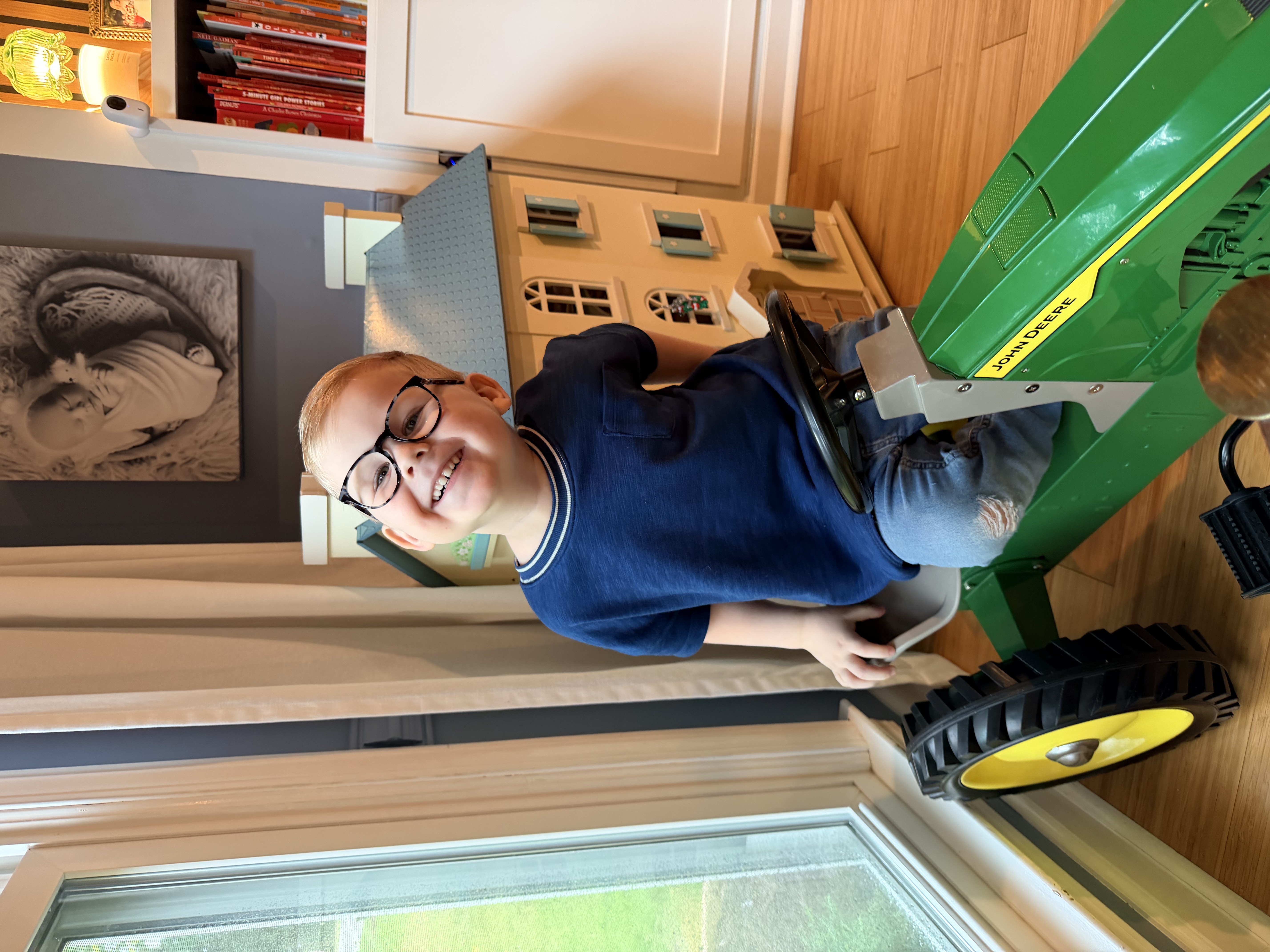 Felix grinning while sitting on his green toy tractor at home